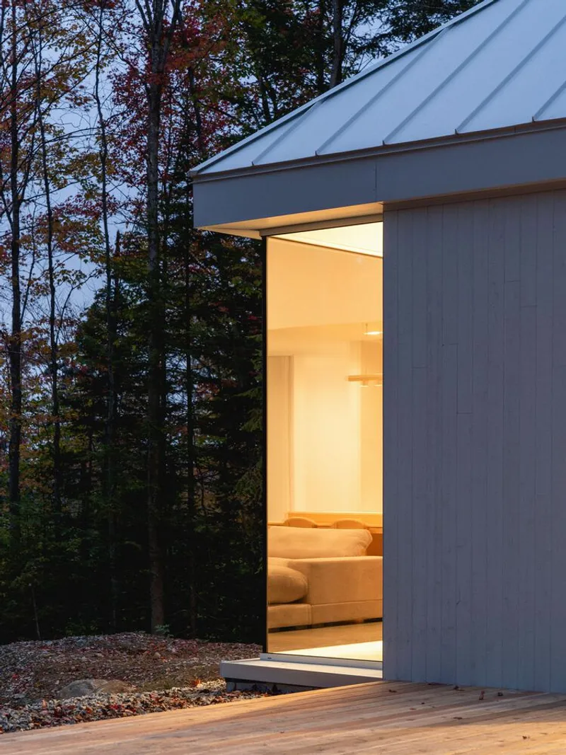 Bedroom window at dusk: warm interior light glowing through glass, cedar deck, gravel ground