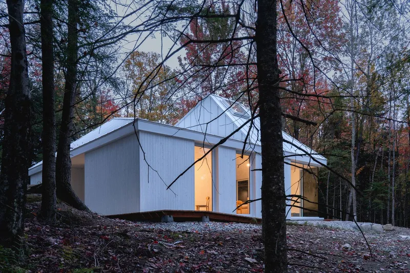 Dusk through autumn trees: white house with warm interior glow, timber deck corner, metal roof catching last light