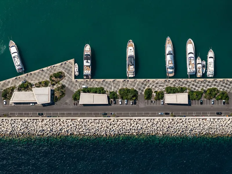 Aerial view of stone breakwater with marina promenade, planted beds and moored yachts in teal water