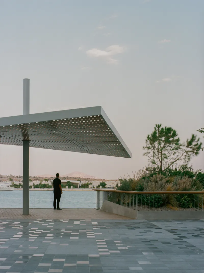 Perforated metal shade canopy over chequered paving with figure overlooking the water at dusk