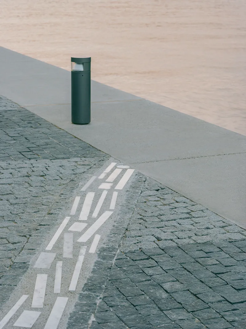 Detail of painted white dashes crossing from stone pavers onto concrete with dark green bollard