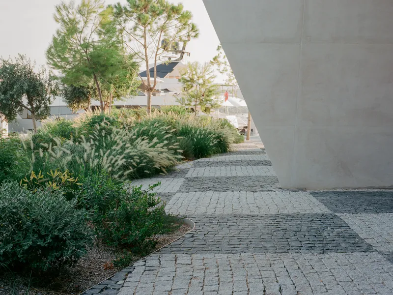 Cobblestone pathway curving beneath a concrete overhang framed by pine trees and native grasses