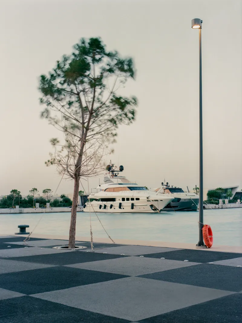 Waterfront plaza with chequered paving, pole-mounted light and red lifesaver beside docked yacht at dusk
