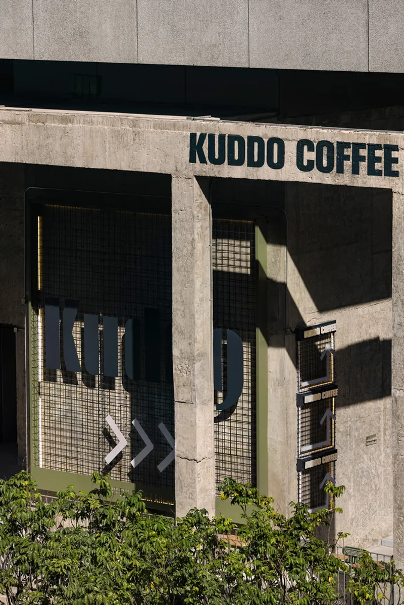 Street facade with exposed concrete frame, mesh-screened openings and signage above low greenery in sunlight