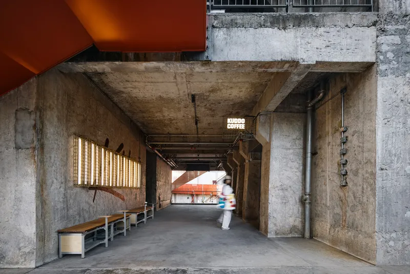 Concrete passage beneath an elevated structure with a timber-slat light installation and benches along the wall