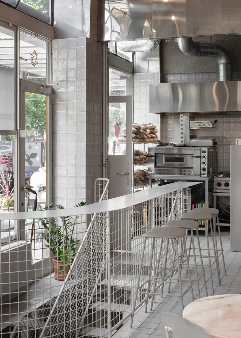 Kitchen through wire mesh: stainless steel bakery equipment visible, stools at the counter, street through the window beyond