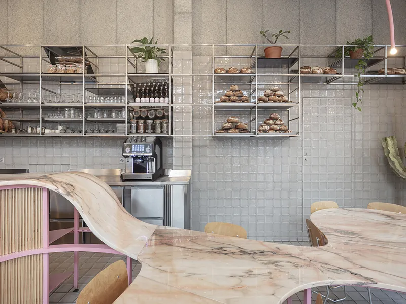 Display wall: metal shelving with pastries, coffee machine, wine bottles, plants, grey tiles, marble counter in foreground