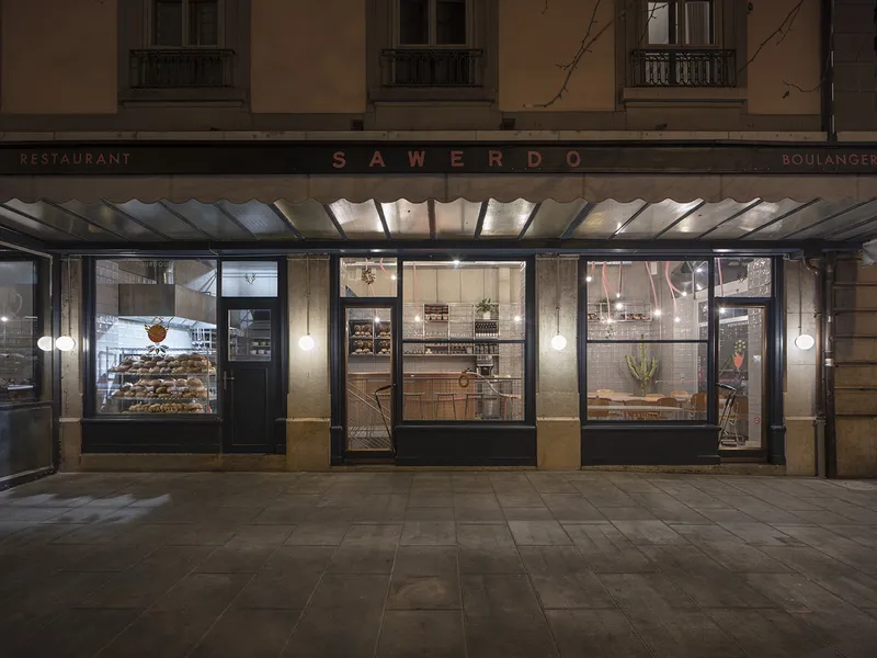 Night exterior: SAWERDO signage on stone facade, black-framed shopfront windows, bakery and cafe glowing warm inside