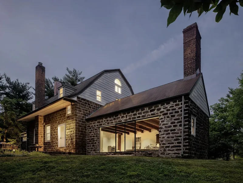 Stone addition with large framed glass opening beside original gabled volumes at twilight