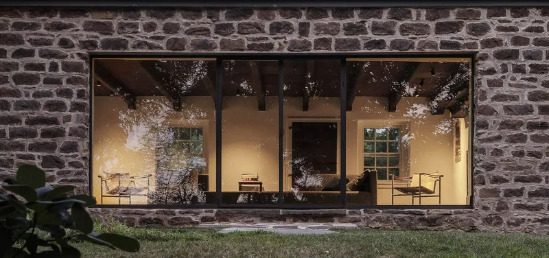Rubble stone wall with large steel-framed windows illuminating the interior at dusk