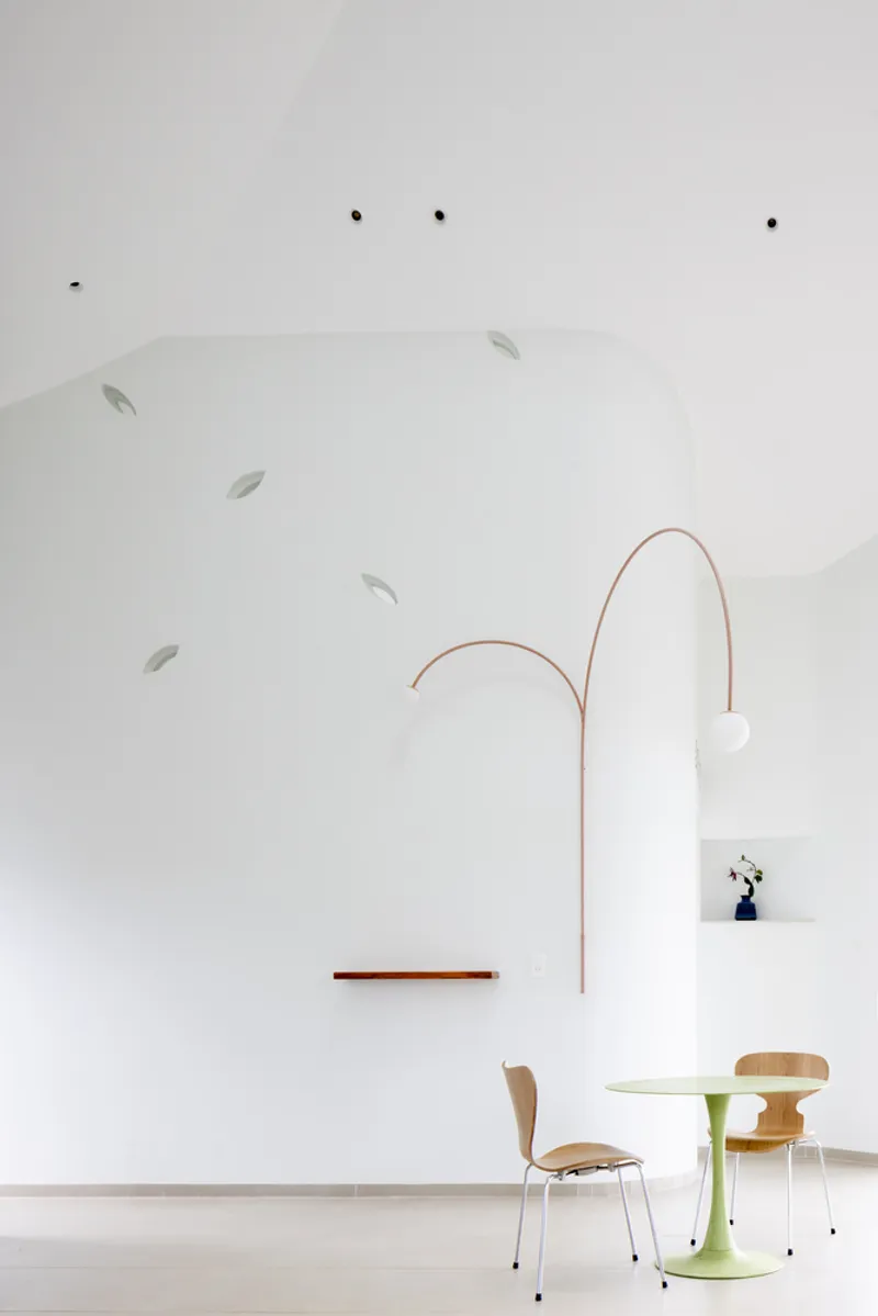Dining area beneath curved ceiling plane with copper arc lamp suspended over plywood chairs and round table