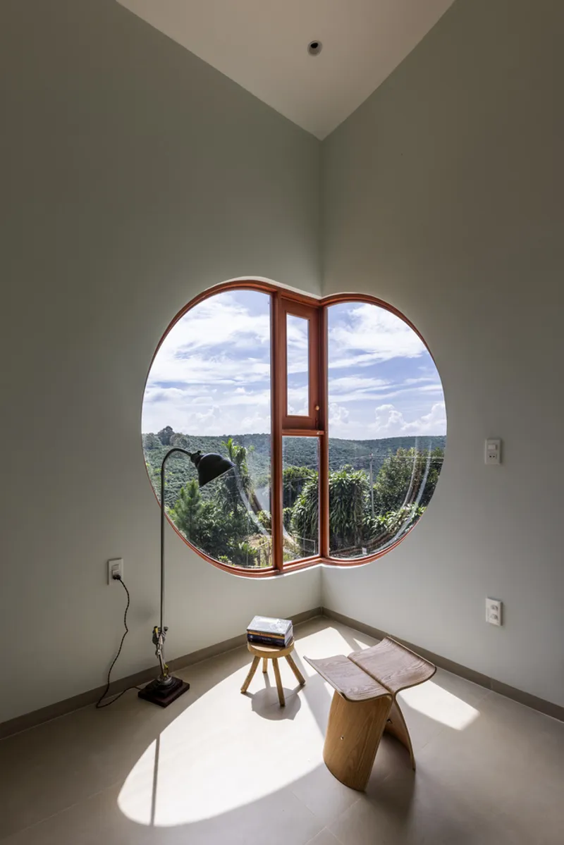 Circular timber window framing rolling green hills under cloudy sky with floor lamp and stool