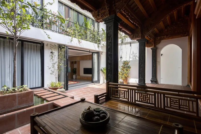 Courtyard from the ancestral hall: dark carved columns, terracotta tiles, low timber tea table, lattice railing, modern house with planted balconies beyond