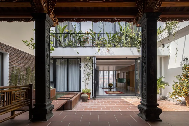 Central courtyard: traditional Vietnamese wooden ancestral hall with carved columns and beams in the foreground, modern house with glass doors behind, terracotta tile floor, bamboo, citrus tree