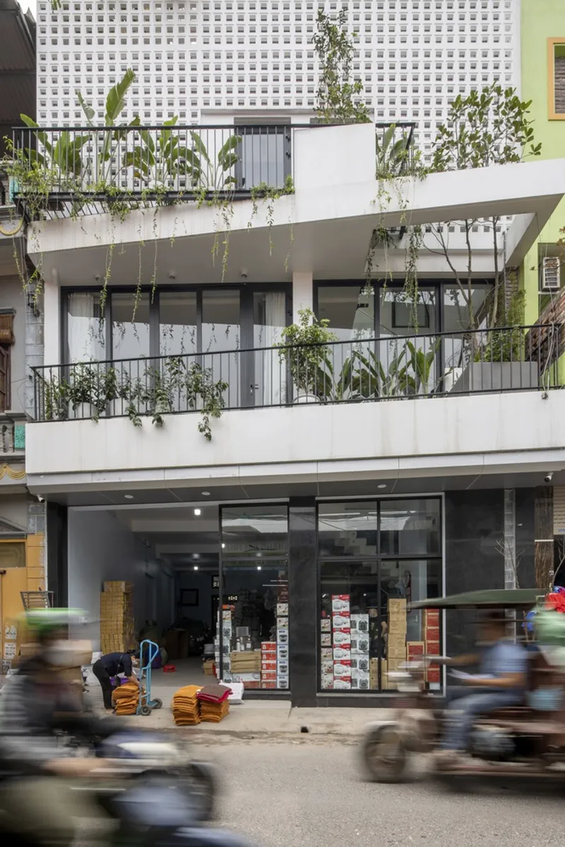 Street facade: white concrete commercial frontage with ground floor shop, upper floors with planted balconies, motorbikes blurring past