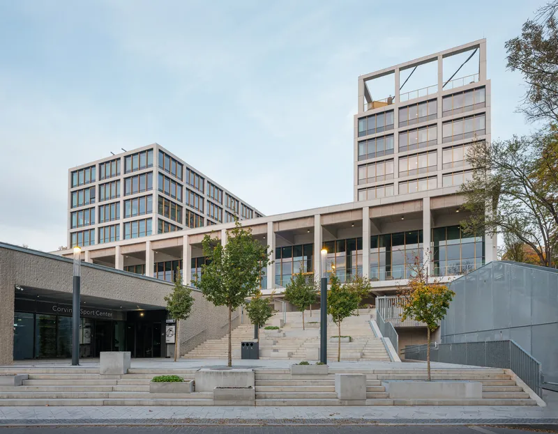 Terraced plaza with young trees leading to two elevated glass and concrete volumes in autumn light