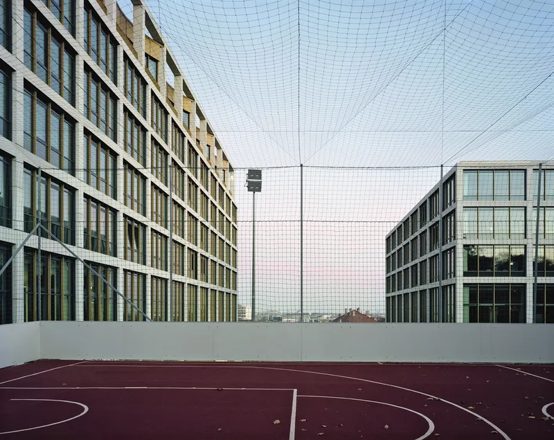 Rooftop basketball court with maroon surface enclosed by netting between two glazed facade blocks at dusk