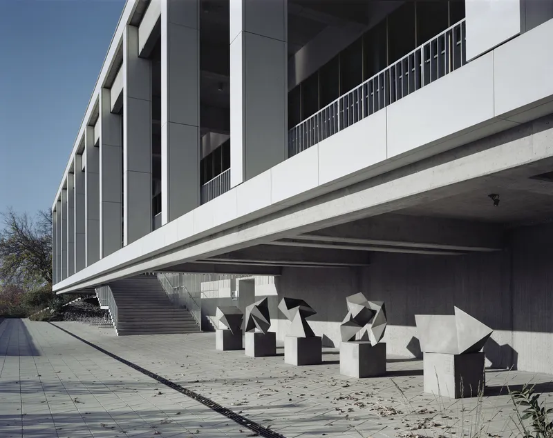 Cantilevered volume with concrete soffit above a paved terrace displaying white and black geometric sculptures