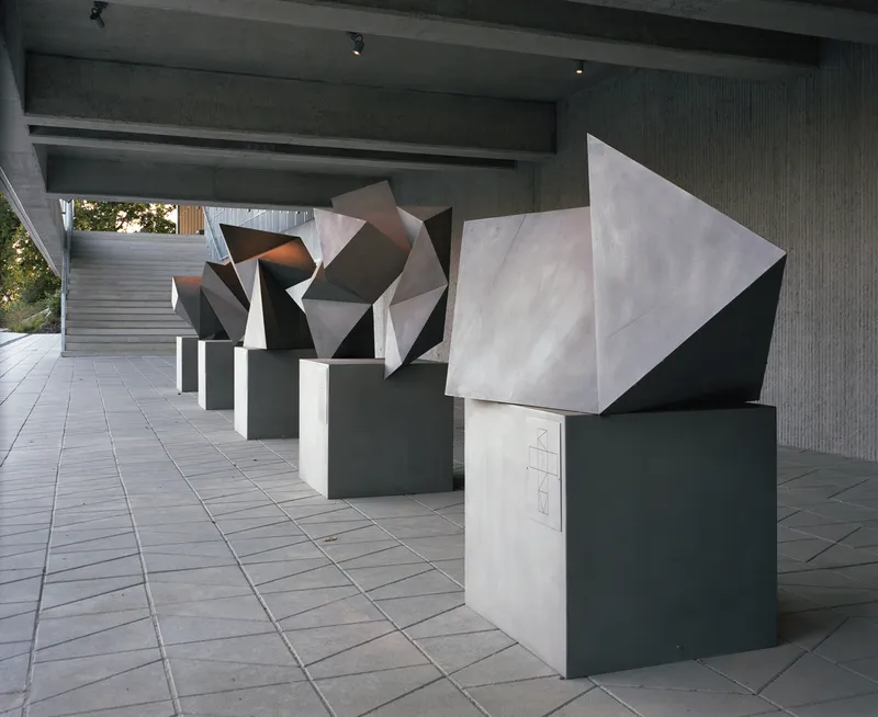 Covered walkway with ribbed concrete ceiling sheltering a row of faceted geometric sculptures on dark plinths