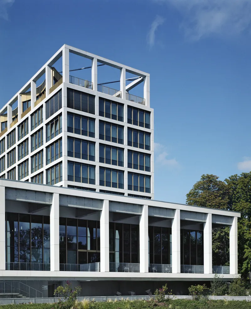 White structural frame building with glass facades rising above tree canopy under blue sky
