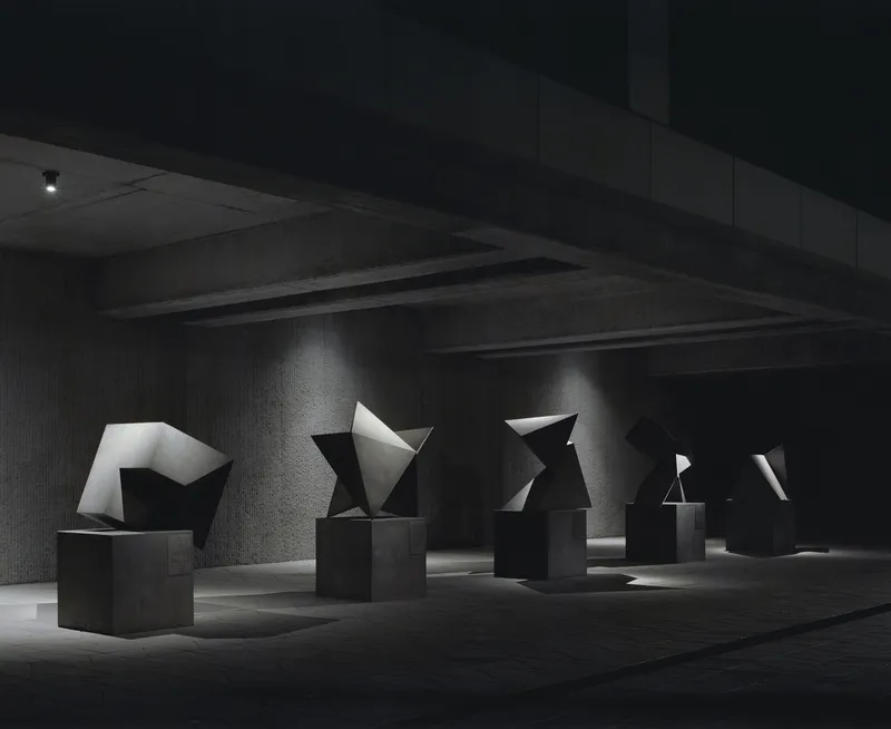 Gallery space with angular geometric sculptures on pedestals beneath exposed concrete beams and dramatic lighting