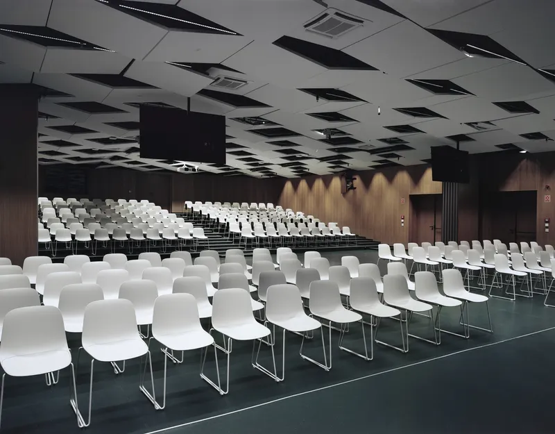Auditorium with rows of white chairs on dark carpet beneath a ceiling of angled black acoustic panels