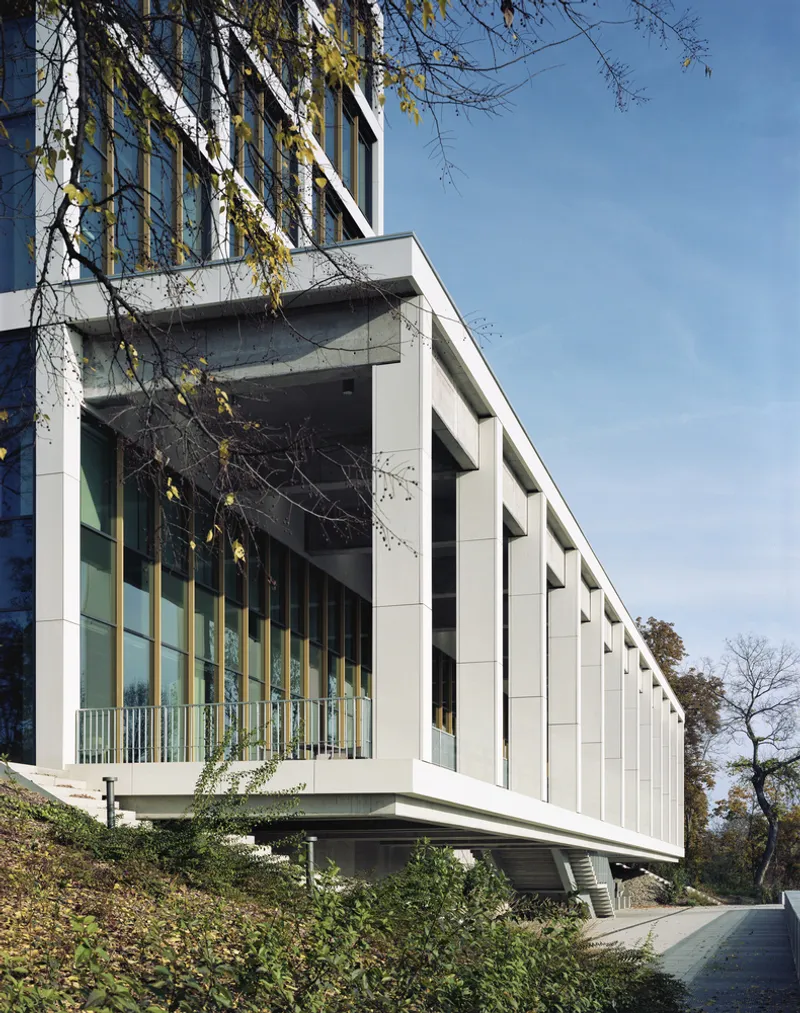 Elevated white concrete volume with vertical glass bays and perforated panels under autumn trees