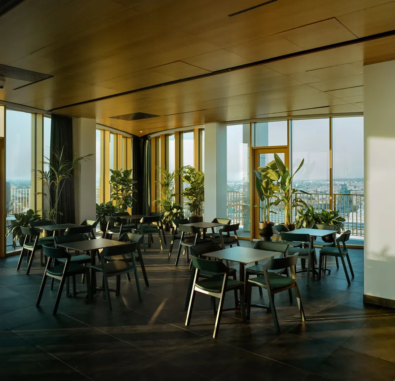 Dining area with timber ceiling panels, dark tables and chairs, and potted palms beside full-height windows