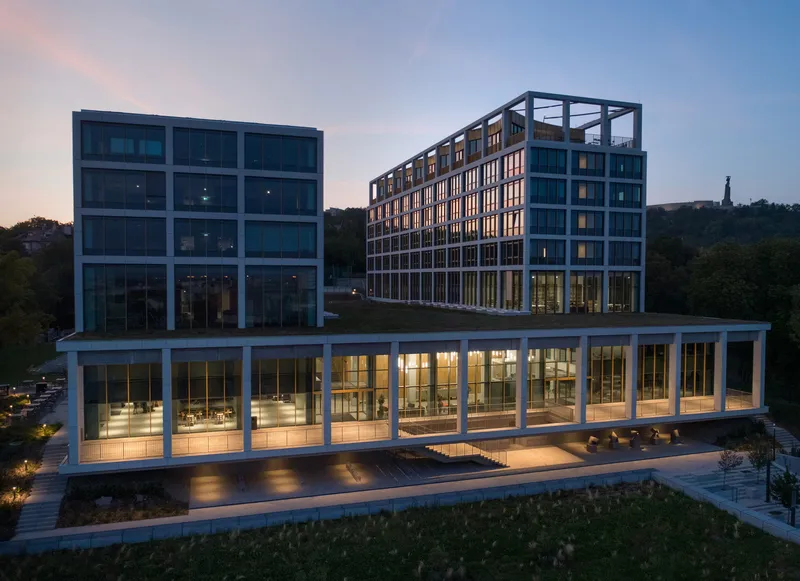 Illuminated glass pavilion base with columned structure beneath two residential blocks at dusk