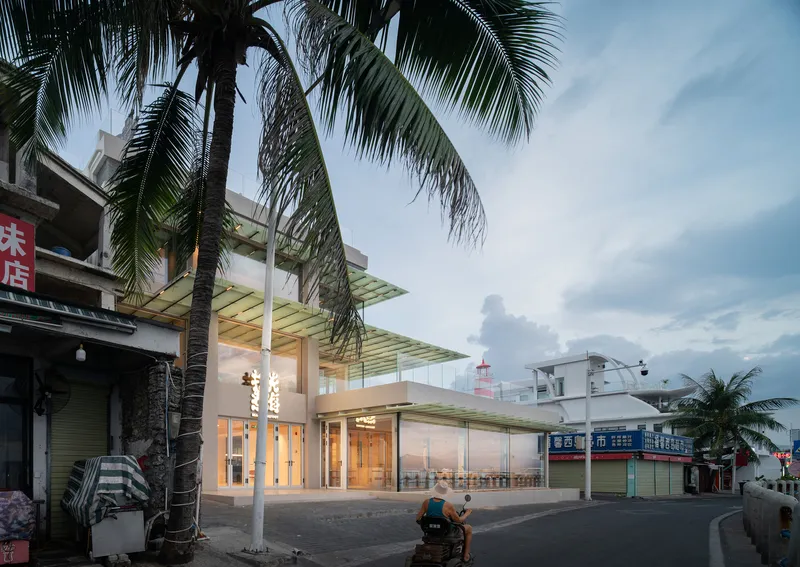 Street view of the glazed pavilion with layered green canopy overhang beneath palm trees at dusk