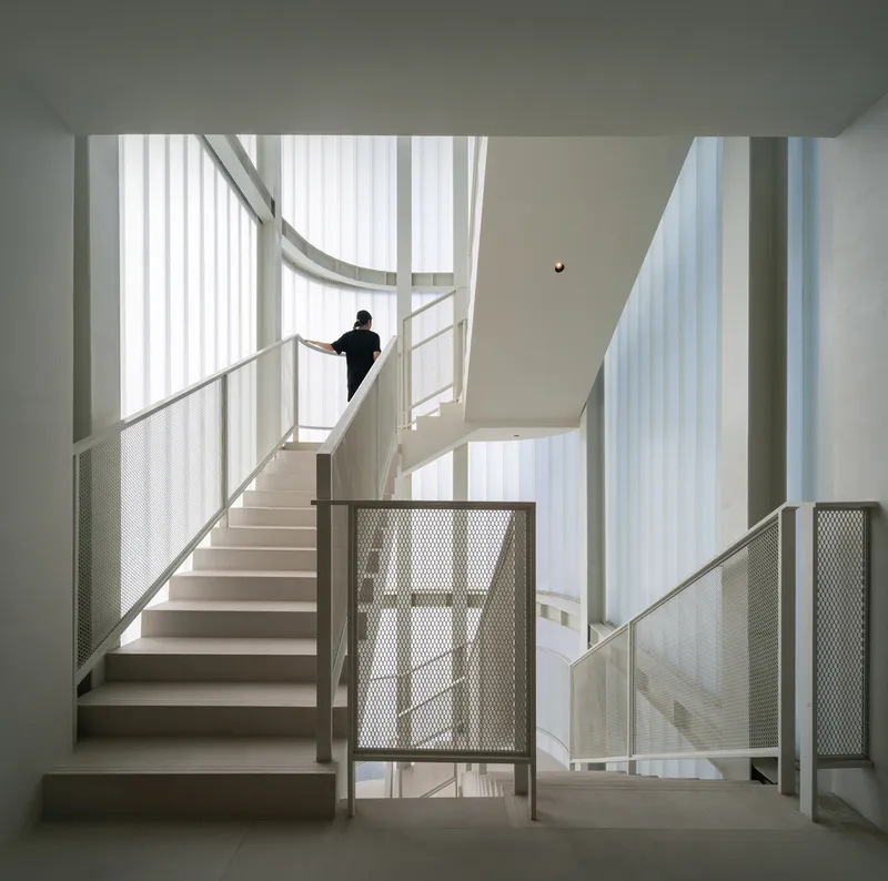 Multi-level stairwell with white metal mesh railings and a figure ascending under diffused natural light
