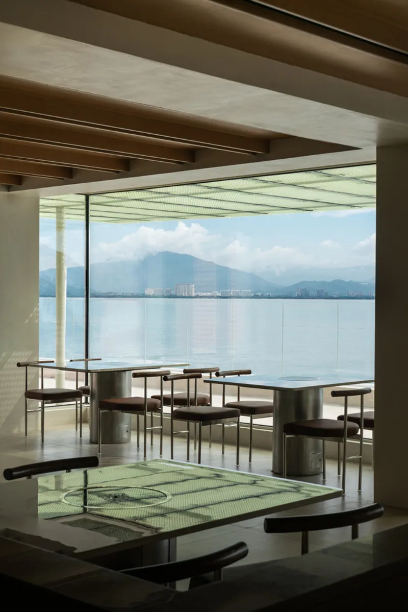 Layered dining spaces with perforated glass floor panels and timber beam ceiling overlooking mountains across the water