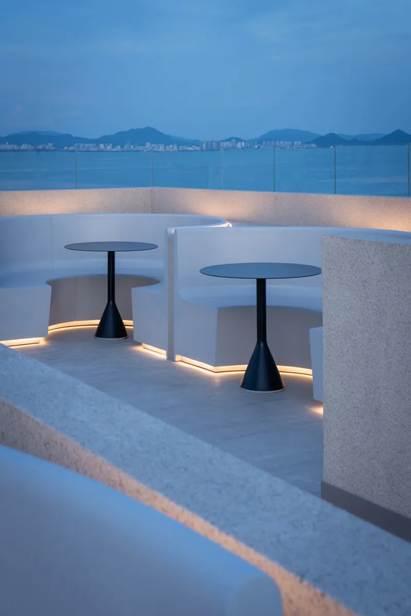 Rooftop terrace with pedestal tables and integrated strip lighting along white planters at dusk overlooking distant mountains