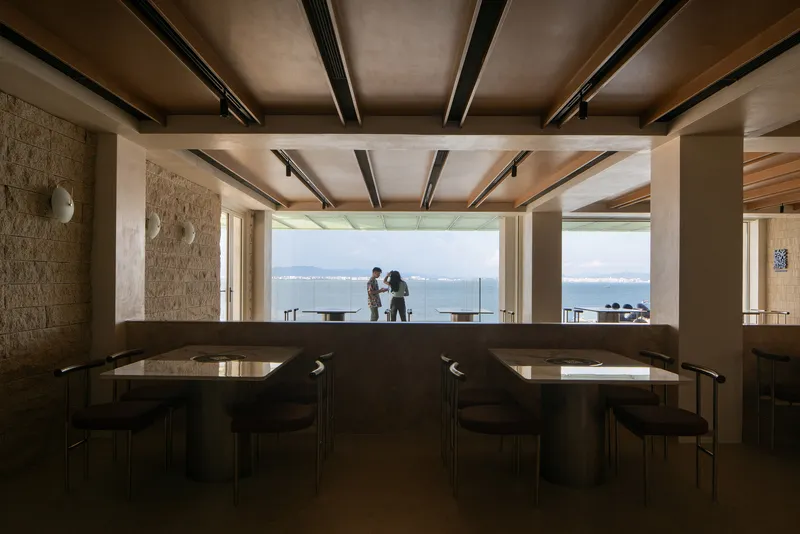 Interior dining area with exposed timber and steel beam ceiling framing a wide harbor view with two figures