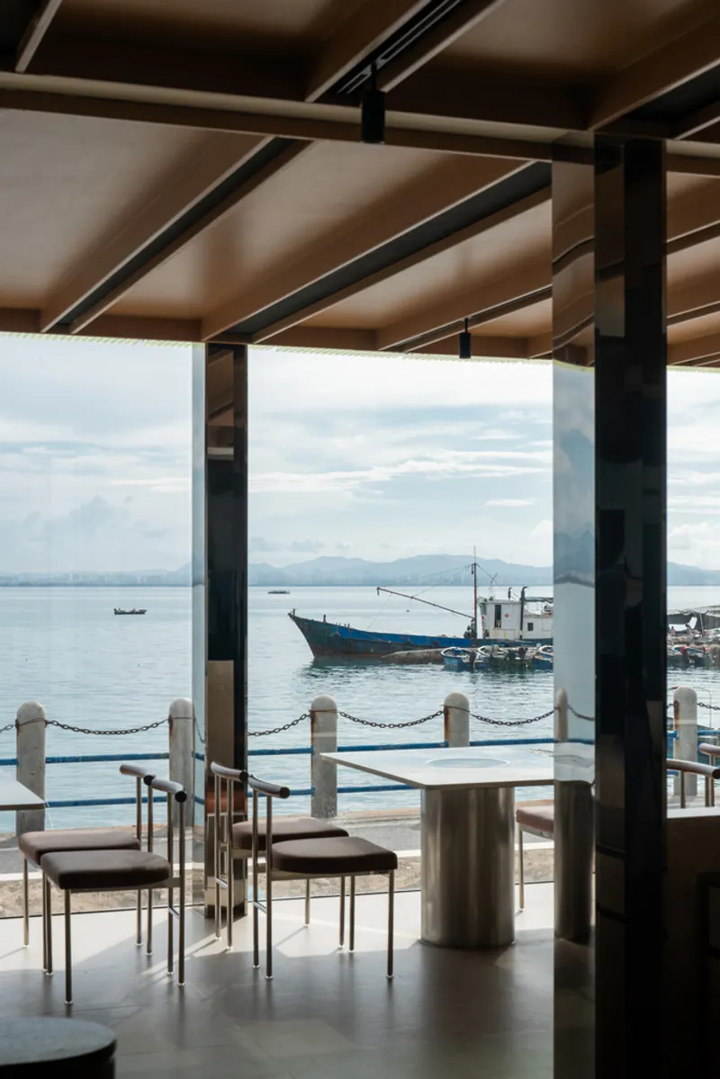 Seating area with timber ceiling structure and chain barrier framing a view of a blue fishing boat at dock