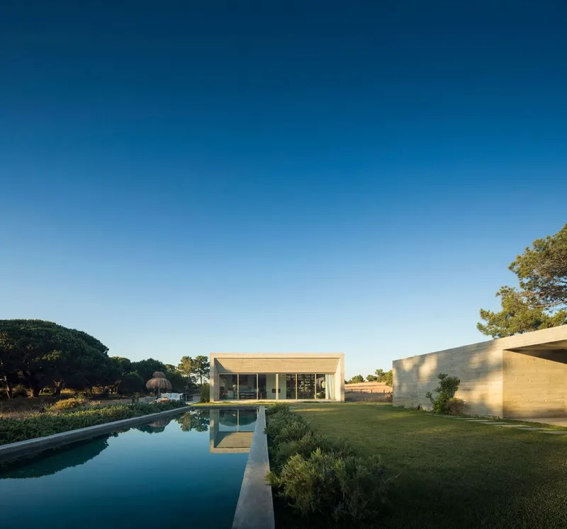 Glass pavilion and board-formed concrete walls beside a lap pool and lawn at twilight