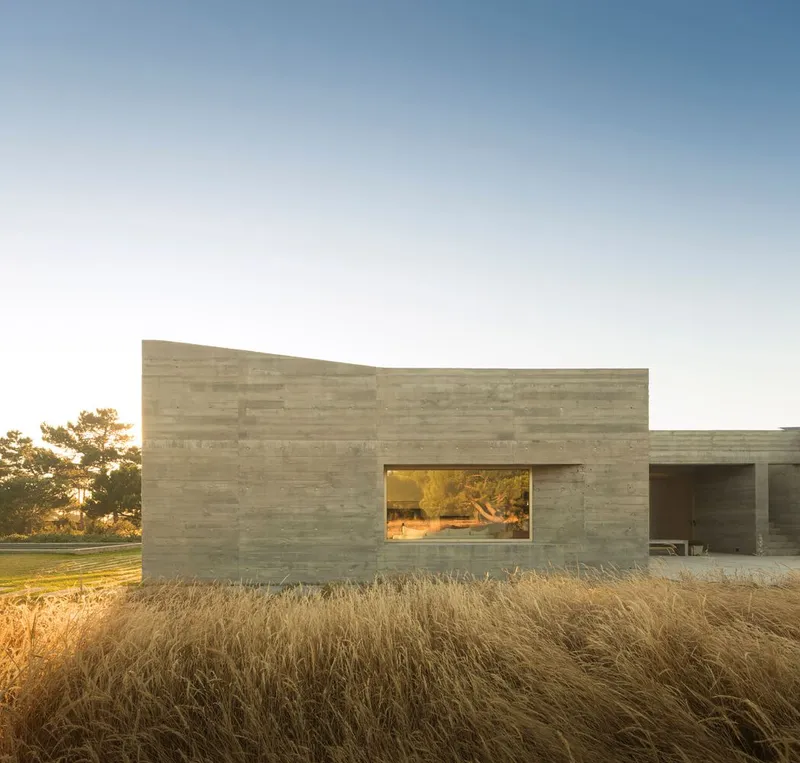 Board-formed concrete volume with a single horizontal window opening above dry grasses at sunset