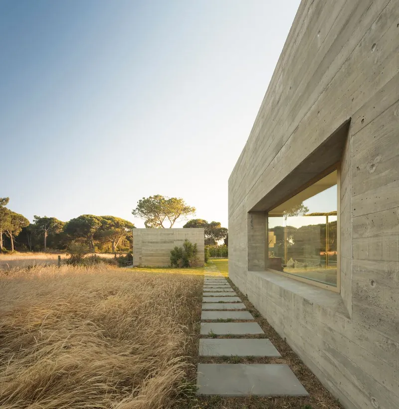 Concrete paver pathway alongside board-formed concrete wall with recessed window and dry grasses