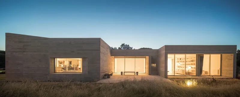 Rammed earth facade with illuminated glazed openings at dusk in a dry grassland