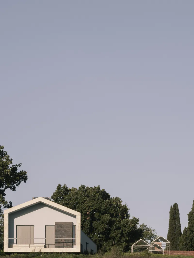 White gabled volume with timber doors beside a skeletal frame structure beneath an overcast sky