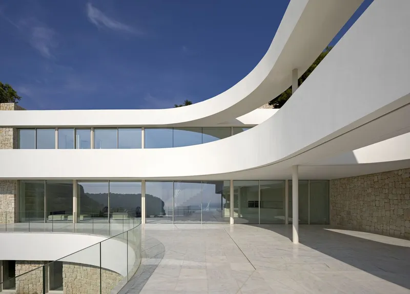Stacked curving terraces with glass balustrades overlooking coastal hills under a clear blue sky