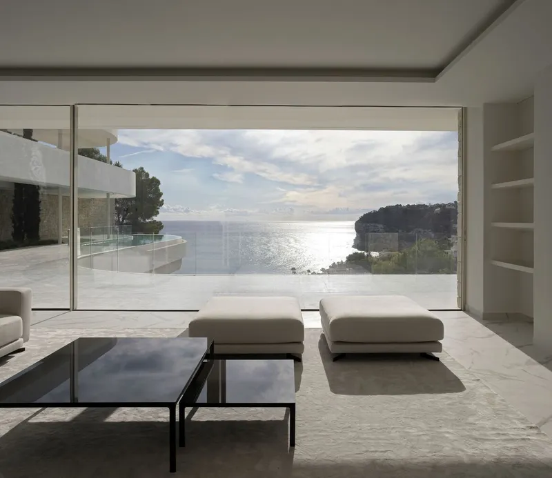 Floor-to-ceiling window framing a coastal view with infinity pool terrace and distant hillside in morning light