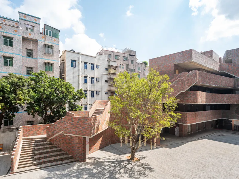 Brick courtyard with cascading staircase and green tree surrounded by mixed residential buildings under midday sun