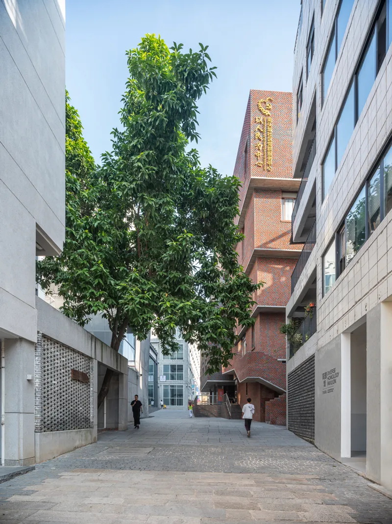 Narrow pedestrian lane between brick and concrete buildings with tall trees and people walking
