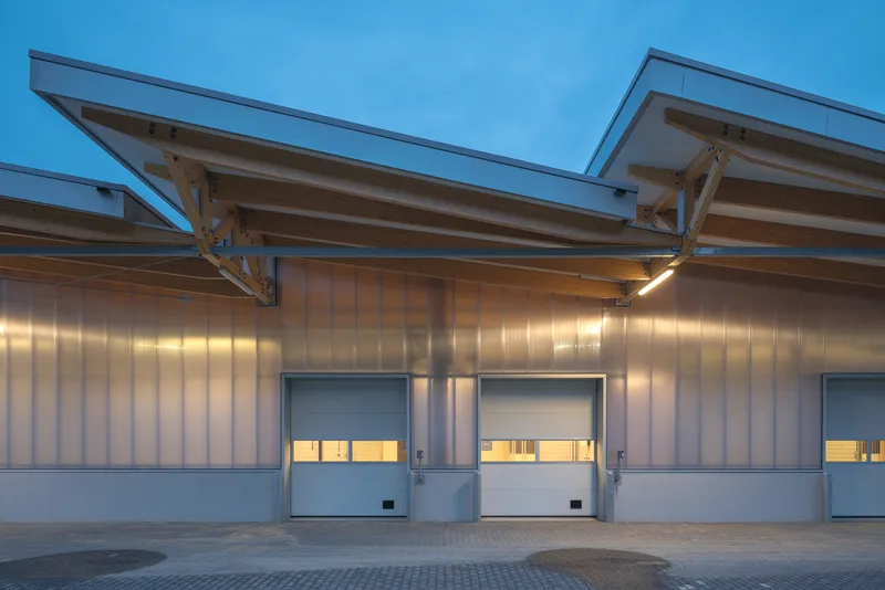 Corrugated metal facade with sawtooth roof trusses and illuminated garage doors at dusk
