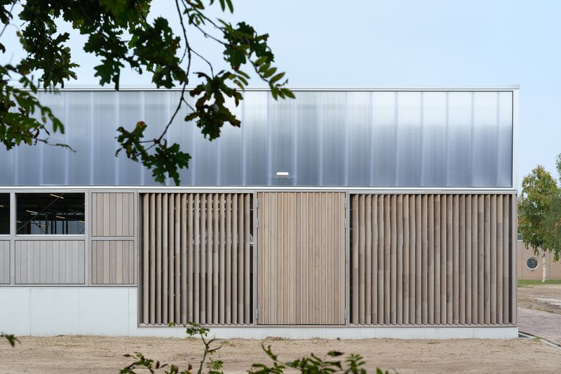 Facade with vertical timber screening below a translucent polycarbonate upper volume framed by tree branches