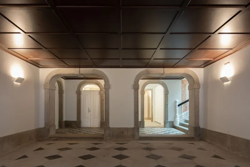 Interior hall with stone arches, diagonal-patterned floor tiles, and coffered wooden ceiling with wall sconces