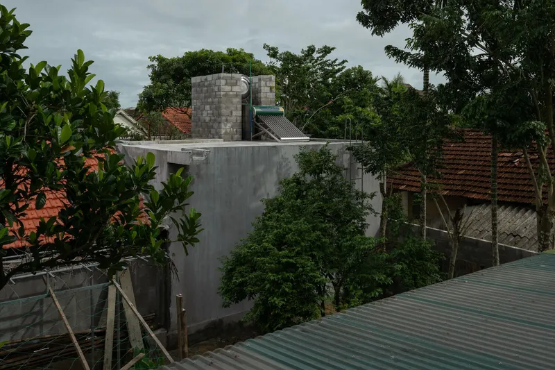 Concrete block volume rising above neighboring tile roofs surrounded by tropical vegetation under overcast skies