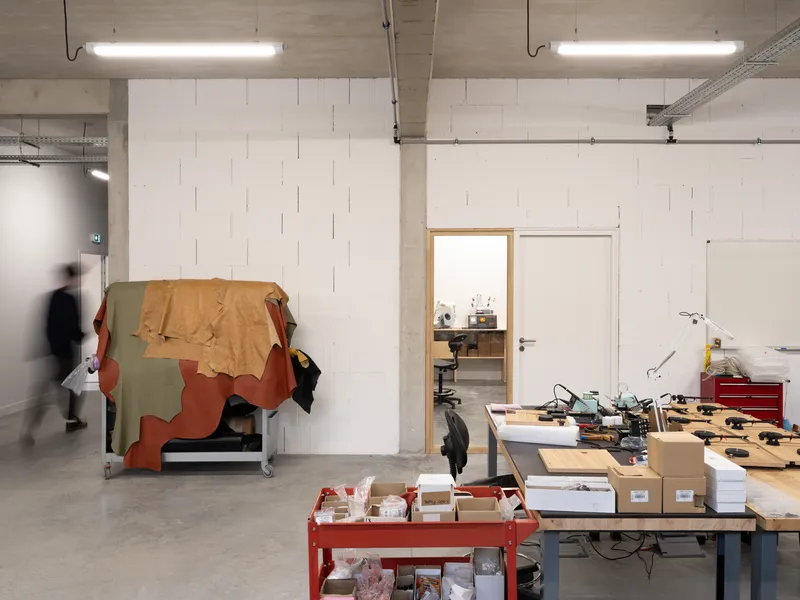 Workshop interior with exposed concrete ceiling, fluorescent tubes, and a person moving past worktables and stored materials