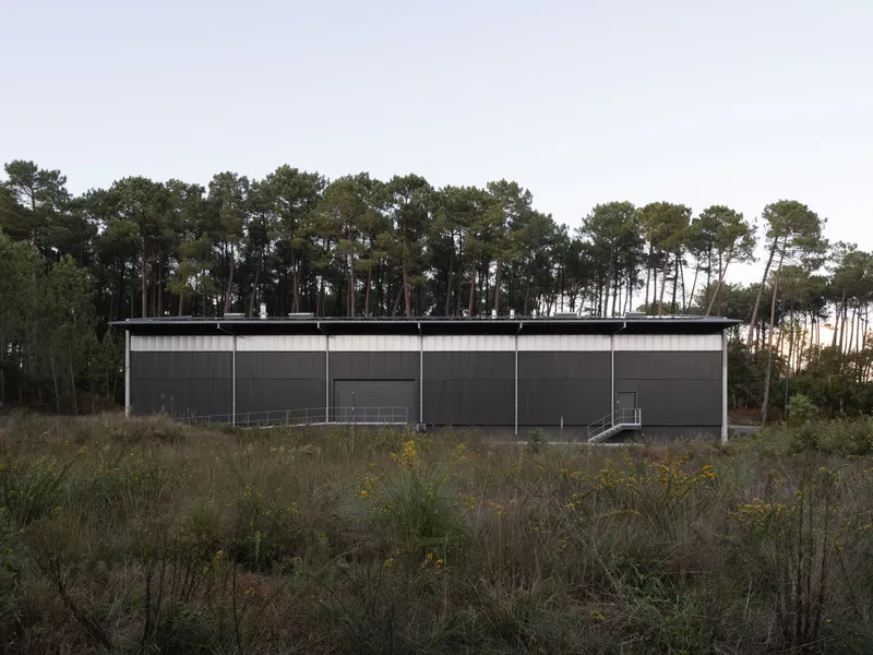 Long corrugated metal facade with flat roof set against pine forest and grassland